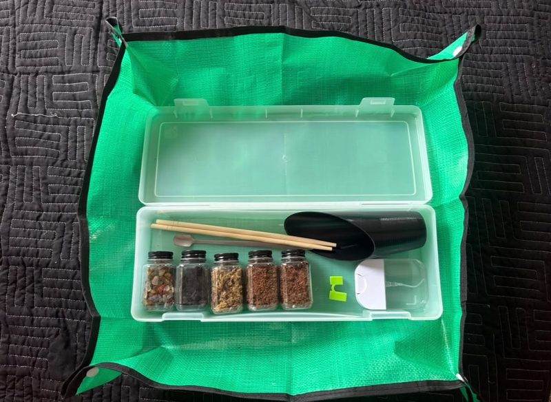 A DIY terrarium kit laid out on a green mess mat: small jars of stone, charcoal, moss and soil, bamboo tools, a scoop and a mini spray bottle.