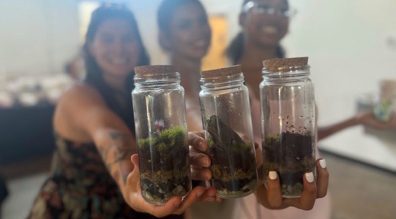 Three smiling people at a workshop holding up the jar terrariums they just built.